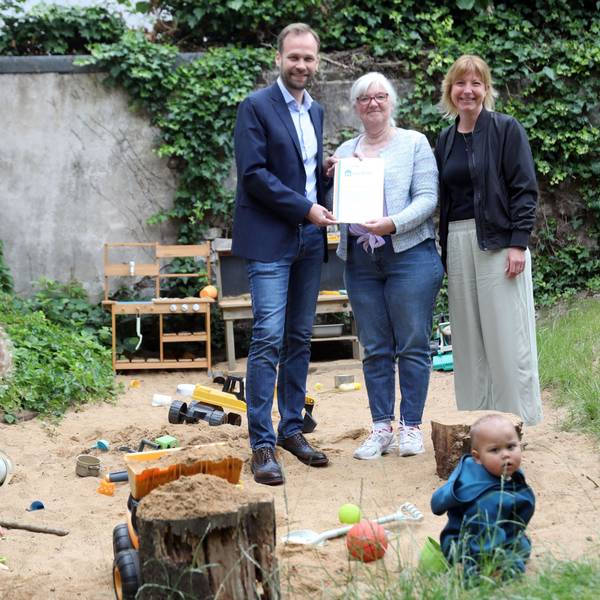 Ein Mann und eine Frau stehen im Hinterhof einer Kindertagesstätte, in dem drei Kinder spielen.