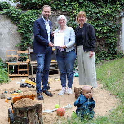 Ein Mann und eine Frau stehen im Hinterhof einer Kindertagesstätte, in dem drei Kinder spielen.