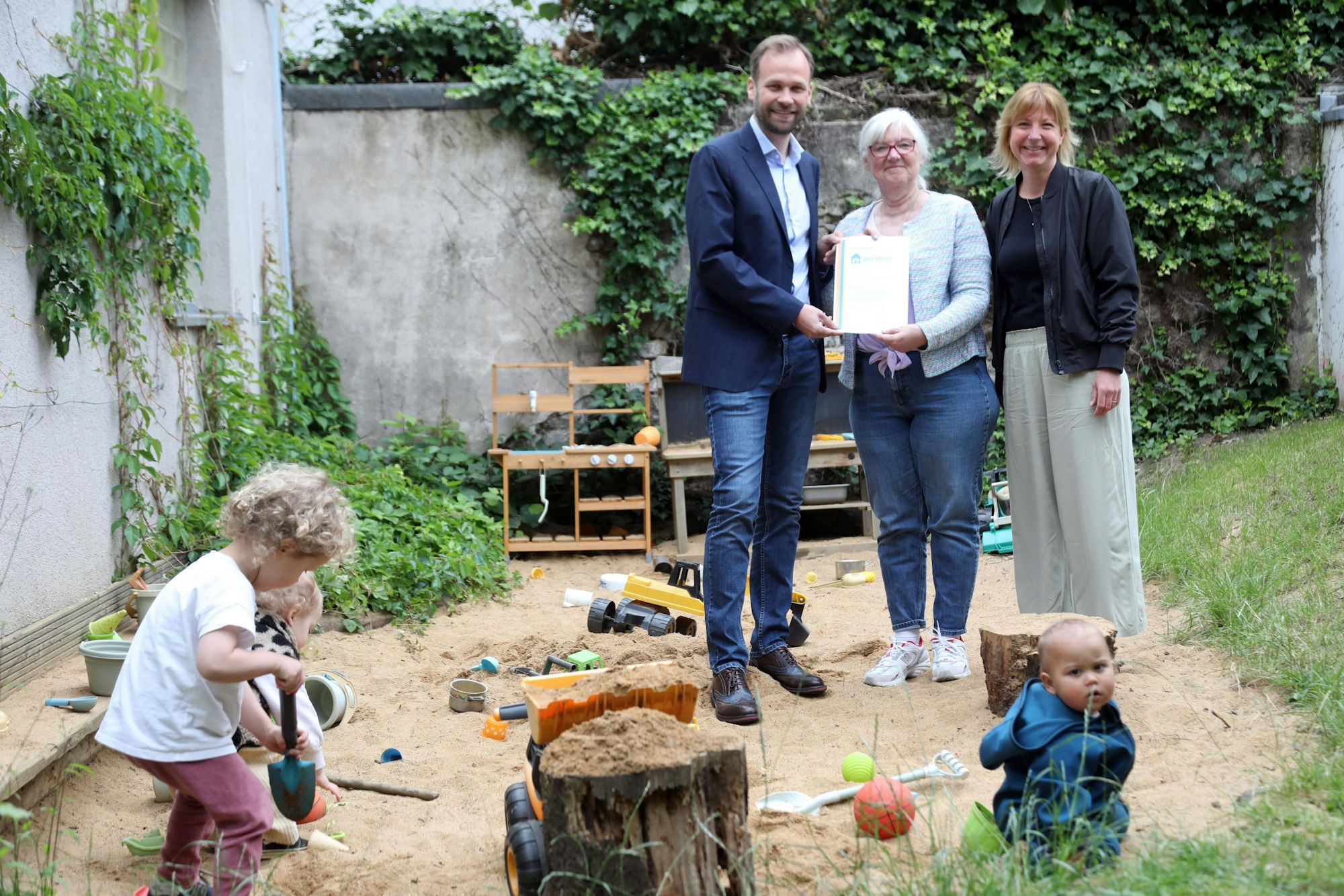 Ein Mann und eine Frau stehen im Hinterhof einer Kindertagesstätte, in dem drei Kinder spielen.