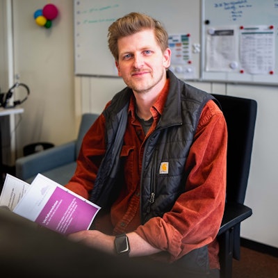 Das Bild zeigt Timo Laas, wie er am Schreibtisch sitzt. In der Hand hält er einen Flyer.