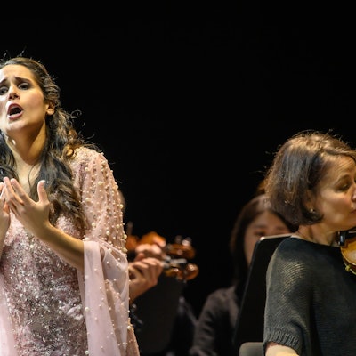 Links im Bild singt Sara Blanch, sie trägt ein rosa Kleid mit Tüll. Neben ihr im Vordergrund eine Violinistin des Gürzenich Orchesters, im Hintergrund verschwommen weitere Musiker.