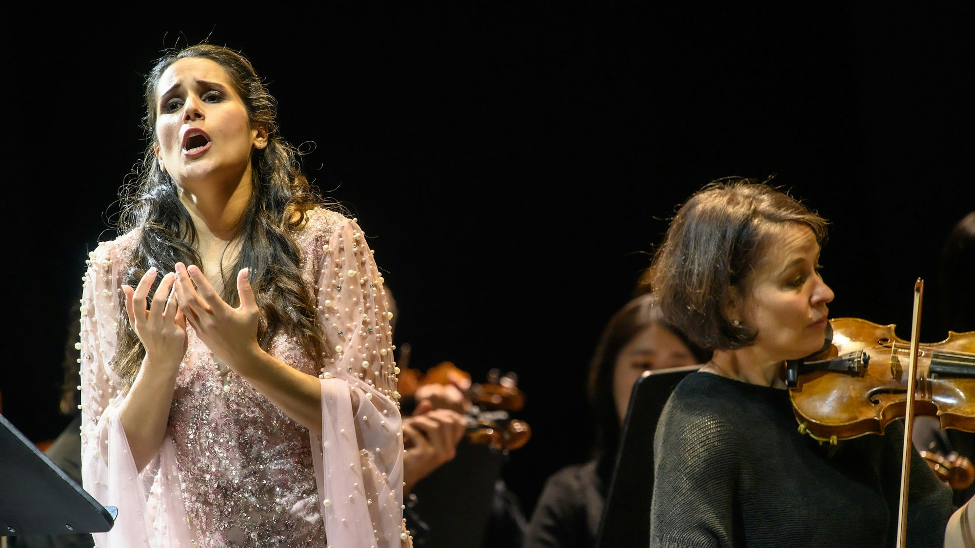 Links im Bild singt Sara Blanch, sie trägt ein rosa Kleid mit Tüll. Neben ihr im Vordergrund eine Violinistin des Gürzenich Orchesters, im Hintergrund verschwommen weitere Musiker.
