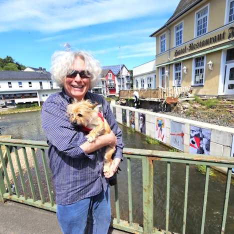 Kurator Jürgen A. Roder trägt seinen Hund auf dem Arm und steht auf einer Brücke vor den Kunstwerken.