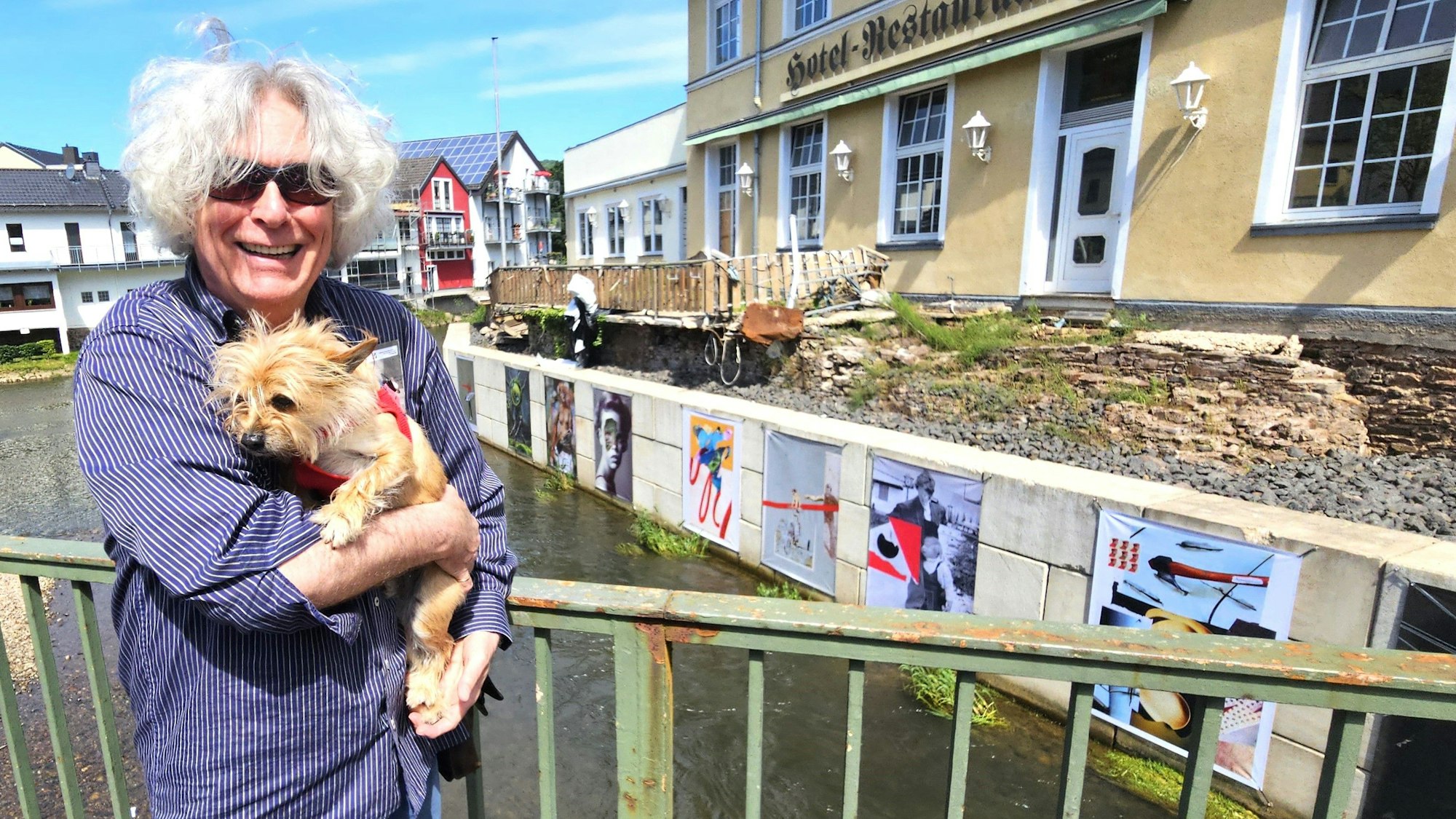 Kurator Jürgen A. Roder trägt seinen Hund auf dem Arm und steht auf einer Brücke vor den Kunstwerken.