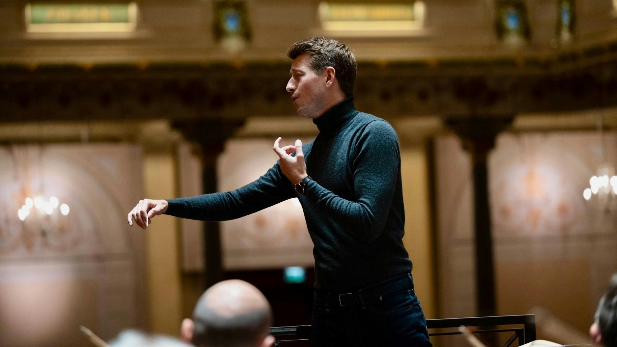 Zu sehen ist Duncan Ward. Er dirigiert mit den Händen. Er trägt einen dunklen Rollkragenpullover und eine Jeans. Im Hintergrund einige Säulen, Kronleuchter und verzierte pfirsichfarbene Wände.