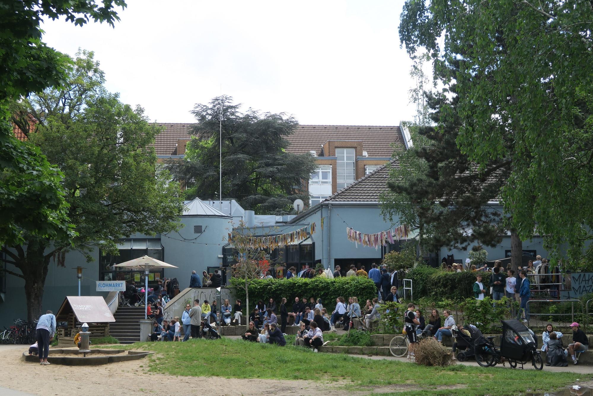Hinter einer Parkfläche mit Spielplatz liegt ein großes Gebäude mit Terrassen.