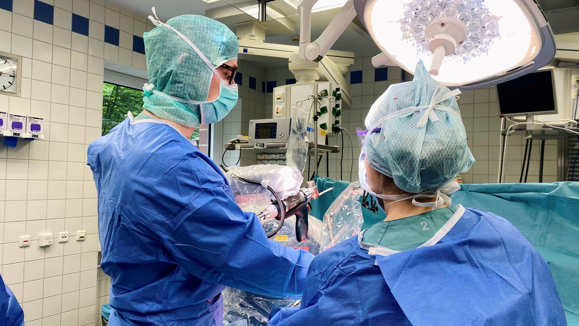 Ein Arzt und eine Ärztin stehen am Operationstisch, auf dem eine Patientin liegt. Zwischen ihnen ist der Bohrarm des Roboters zu sehen.
