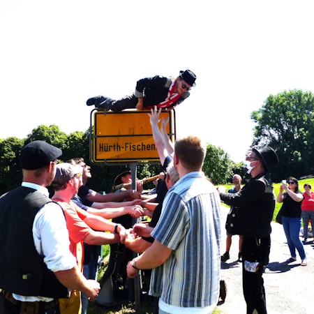 Ein junger Mann mit Hut und Schal klettert über das Ortseingangsschild von Hürth-Fischenich. Ein Empfangskomitee will ihn auffangen.
