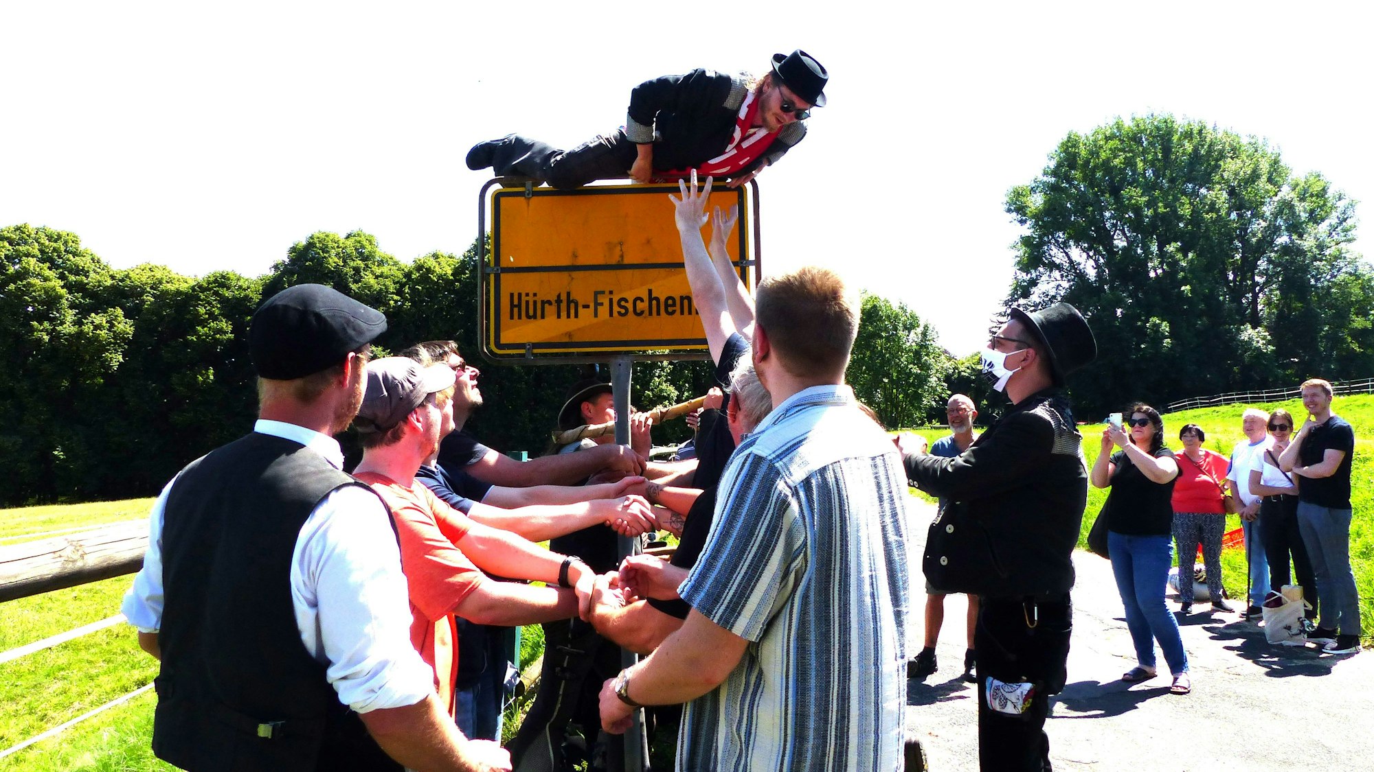 Ein junger Mann mit Hut und Schal klettert über das Ortseingangsschild von Hürth-Fischenich. Ein Empfangskomitee will ihn auffangen.