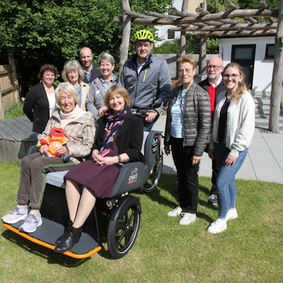 Zwei Frauen sitzen in einer Fahrradrikscha, eine Menschengruppe umringt sie. Einer von ihnen trägt einen Fahrradhelm.