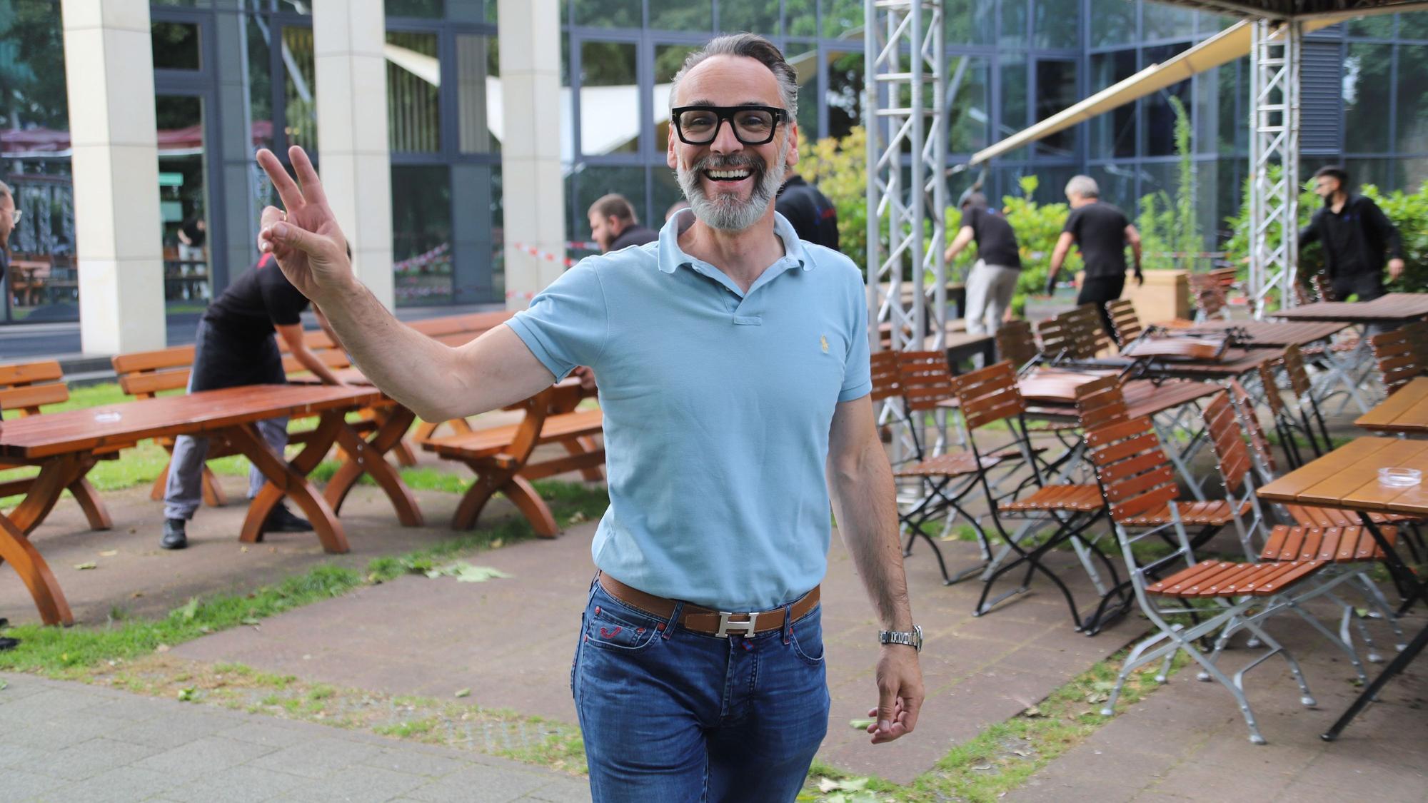 Ein Mann mit Jeans und Brille steht vor Biertischen und hebt zwei Finger.