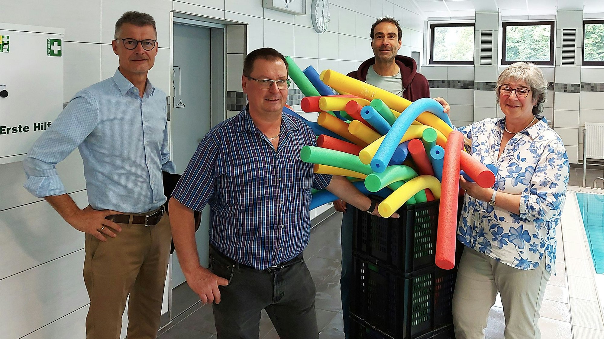 Drei Männer und eine Frau, Jan Lembach, Norbert Jenniches, Elmar Malburg und Marita Schramm, stehen in einem Hallenbad an einem Korb, der mit bunten Poolnudeln gefüllt ist.