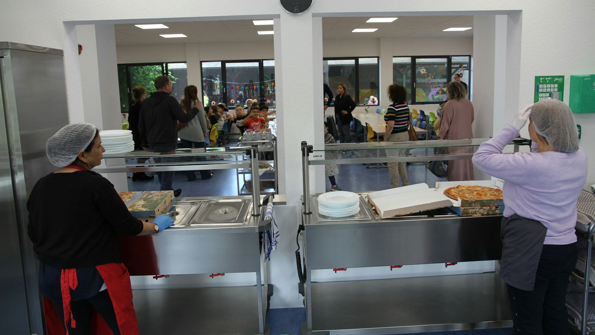 Zwei Frauen mit Schutzhauben stehen an einer Essensausgabe, im Hintergrund ein Speiseraum