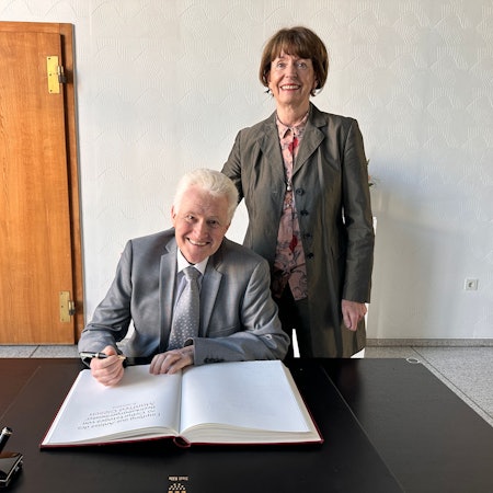 Ein grauhaariger Mann in Anzug und Krawatte sitzt an einem Tisch, Füller in der Hand, vor einem großen Buch. Neben ihm steht eine Frau im Anzug.