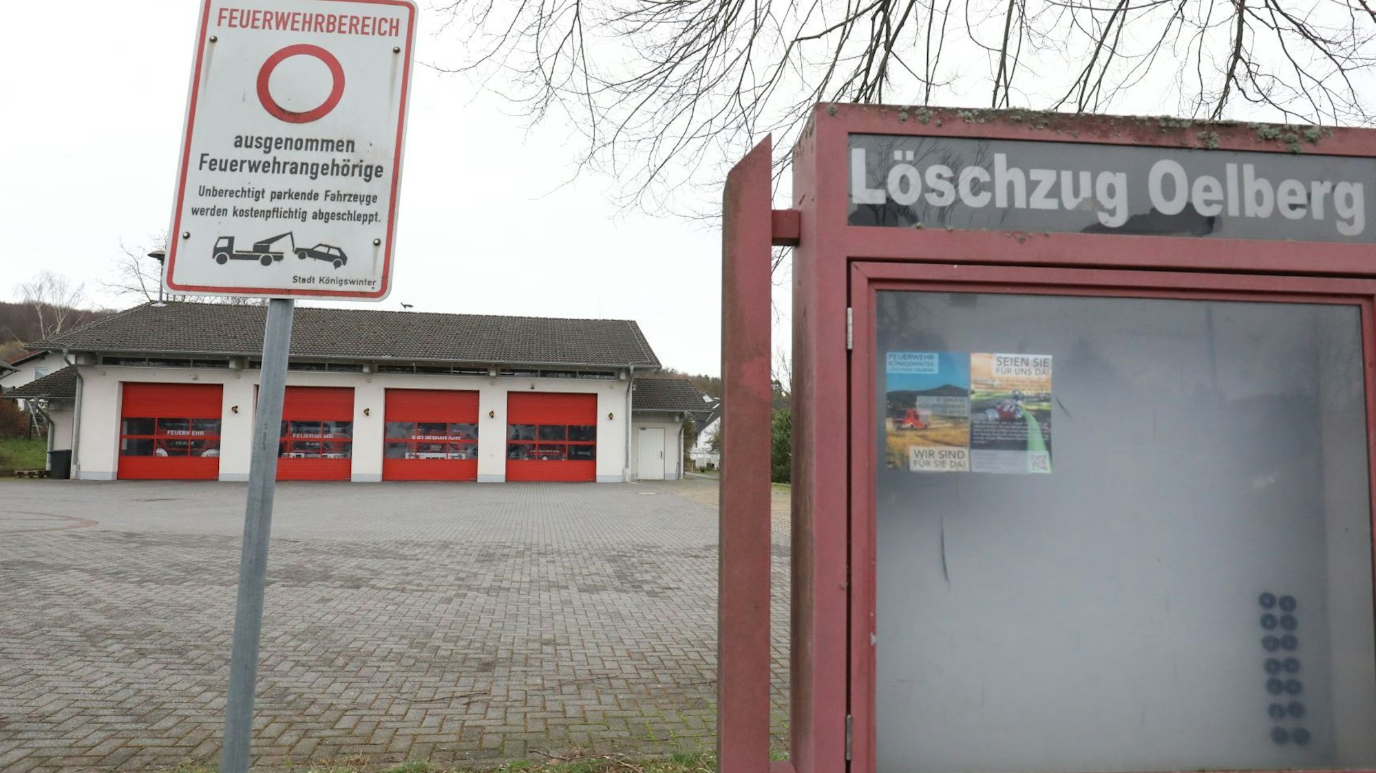 Ein leerer Schaukasten mit der Aufschrift Löschzug Oelberg, im Hintergrund das Gerätehaus mit vier roten Rolltoren.