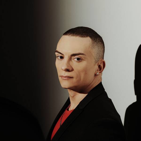 Cameron Carpenter schaut seitlich in die Kamera. Er trägt ein schwarzes Sakko und darunter ein rotes T-Shirt. Er trägt auch einen Ohrring. Im Hintergrund eine weiße Wand, auf die sein Schatten fällt.