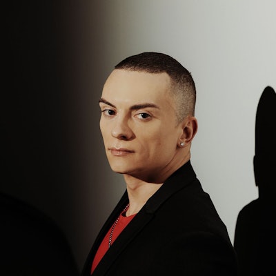 Cameron Carpenter schaut seitlich in die Kamera. Er trägt ein schwarzes Sakko und darunter ein rotes T-Shirt. Er trägt auch einen Ohrring. Im Hintergrund eine weiße Wand, auf die sein Schatten fällt.