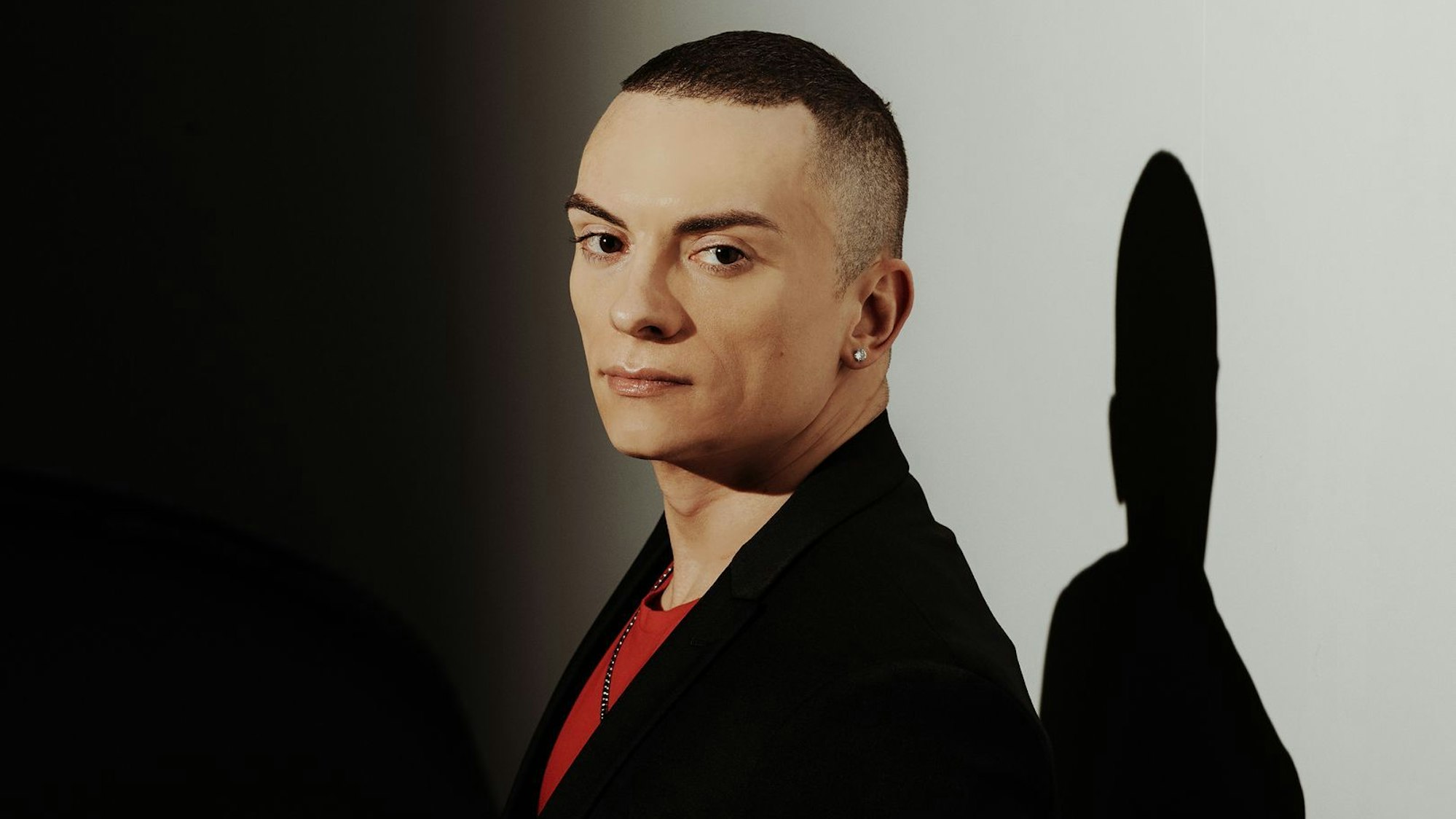 Cameron Carpenter schaut seitlich in die Kamera. Er trägt ein schwarzes Sakko und darunter ein rotes T-Shirt. Er trägt auch einen Ohrring. Im Hintergrund eine weiße Wand, auf die sein Schatten fällt.