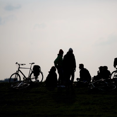 Junge Menschen stehen und sitzen auf dem Boden, Fahrräder neben sich. Das Foto ist gegen die Sonne aufgenommen, ihre Silhouetten sehen wie Schattenrisse aus.