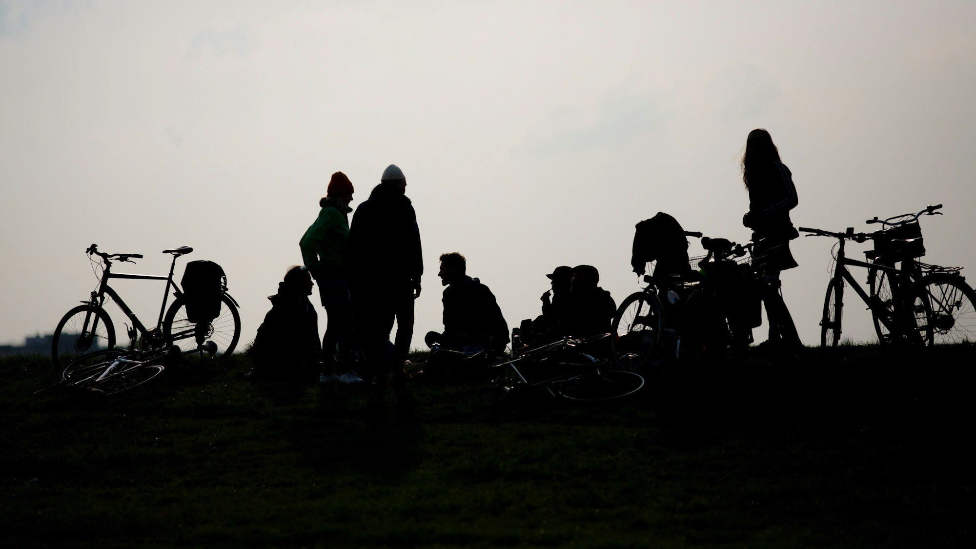 Junge Menschen stehen und sitzen auf dem Boden, Fahrräder neben sich. Das Foto ist gegen die Sonne aufgenommen, ihre Silhouetten sehen wie Schattenrisse aus.