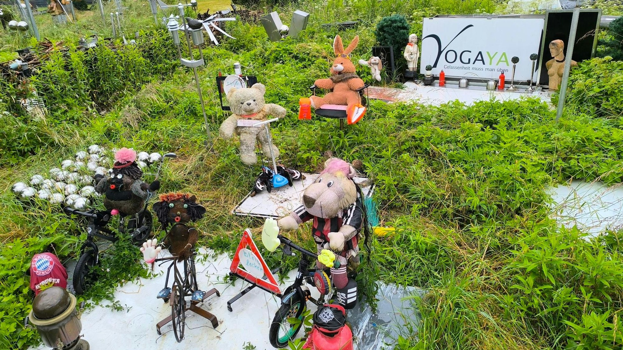 In diesem verwilderten Garten inmitten einer Wohnsiedlung in Leverkusen-Schlebusch sind einige Skulpturen und Figuren zu sehen. Oben rechts im Bild ist ein Schild mit der Aufschrift „Yogaya Gelassenheit muss man üben“ zu sehen.