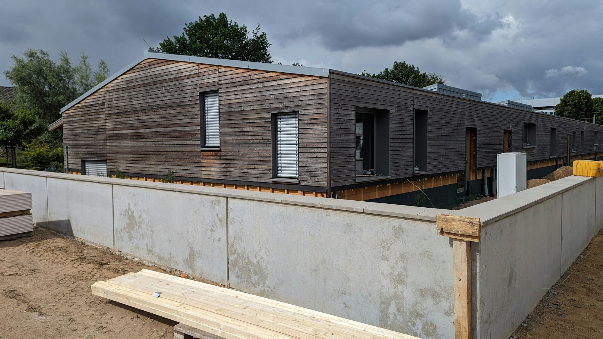 Auf dem Foto ist eine Kita in Holzbauweise zu sehen. Um sie herum wird eine Mauer gegen Überflutungen gebaut.
