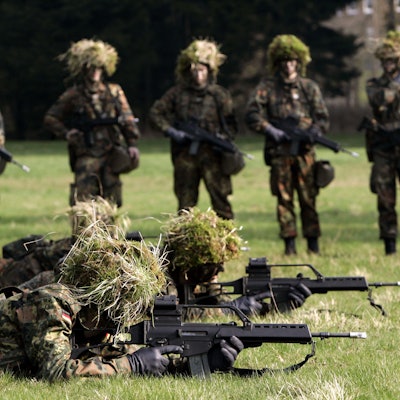 Hessen, Schwarzenborn: Mit Blattwerk und Gras getarnte Wehrpflichtige von den Panzergrenadieren liegen mit Gewehren des Typs G36 auf dem Bauch in Deckung, im Hintergrund werden sie von ihren Kameraden beobachtet. (Archivbild)
