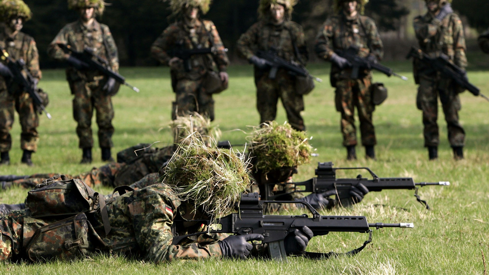 Hessen, Schwarzenborn: Mit Blattwerk und Gras getarnte Wehrpflichtige von den Panzergrenadieren liegen mit Gewehren des Typs G36 auf dem Bauch in Deckung, im Hintergrund werden sie von ihren Kameraden beobachtet. (Archivbild)