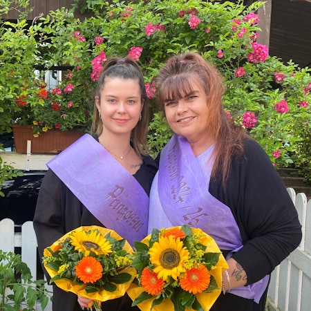Zwei Frauen mit Schärpen und Blumen stehen vor einem Zaun.