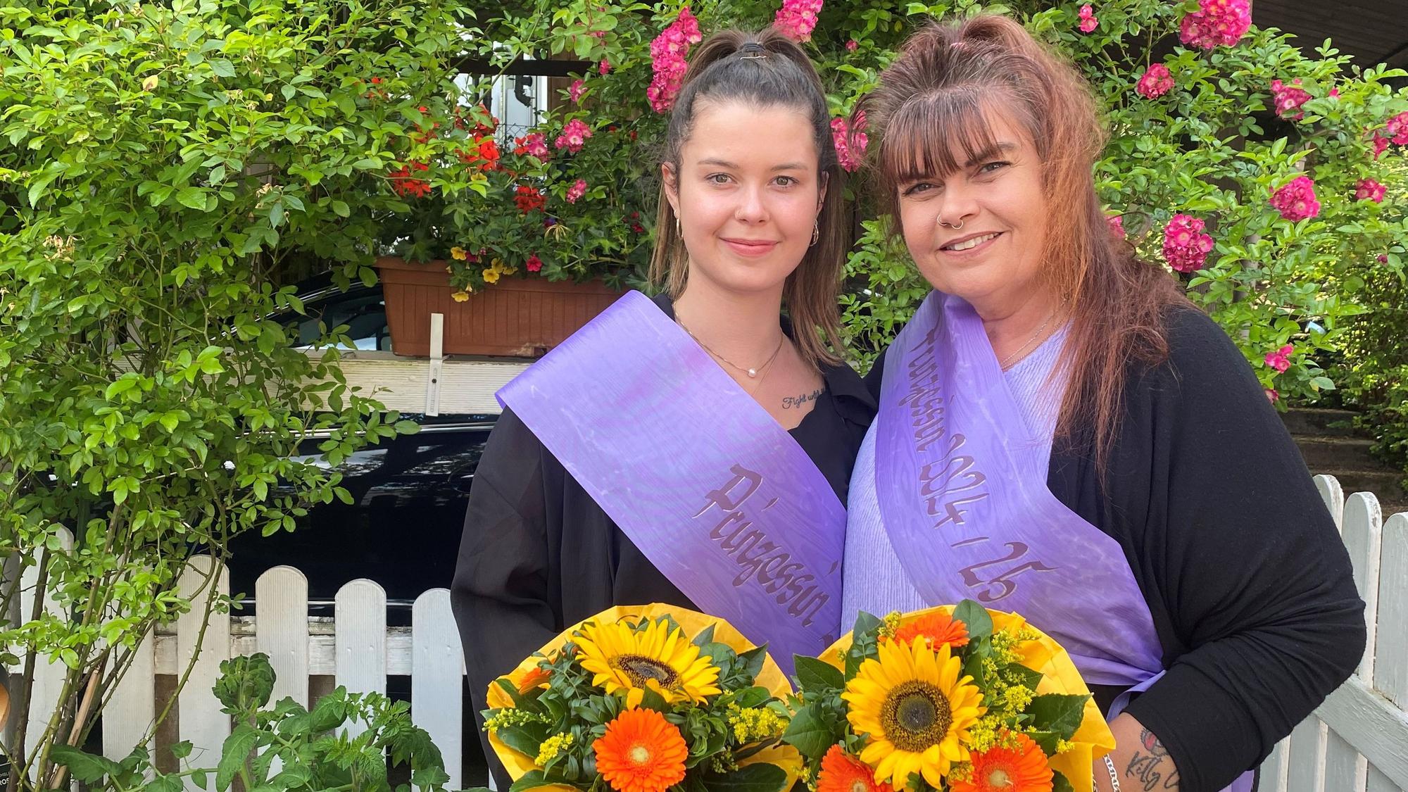 Zwei Frauen mit Schärpen und Blumen stehen vor einem Zaun.