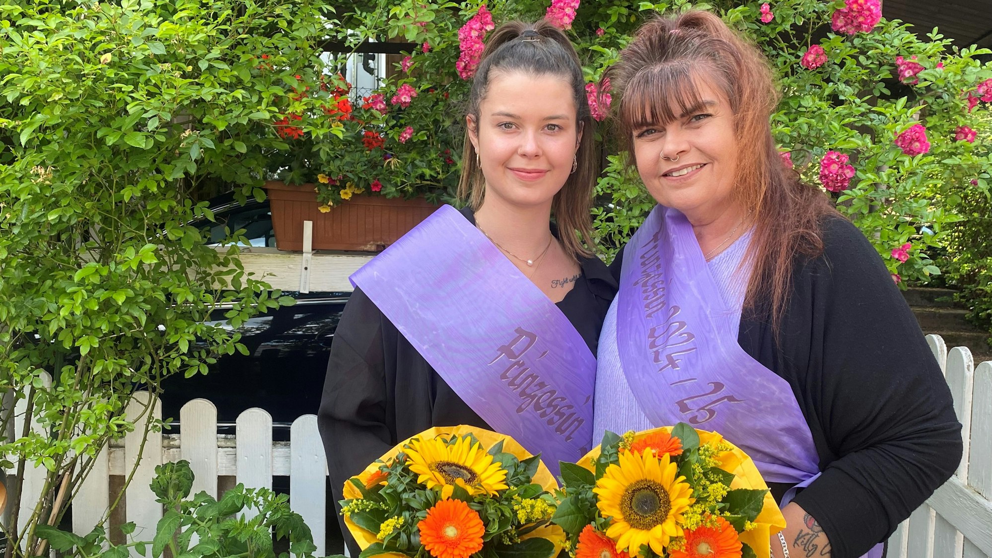 Zwei Frauen mit Schärpen und Blumen stehen vor einem Zaun.