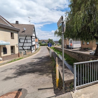 Im Bereich der Rehgasse in Kommern verläuft der Bleibach zwischen alten Ufermauern. Vorne ist das Geländer einer Brücke zu sehen.