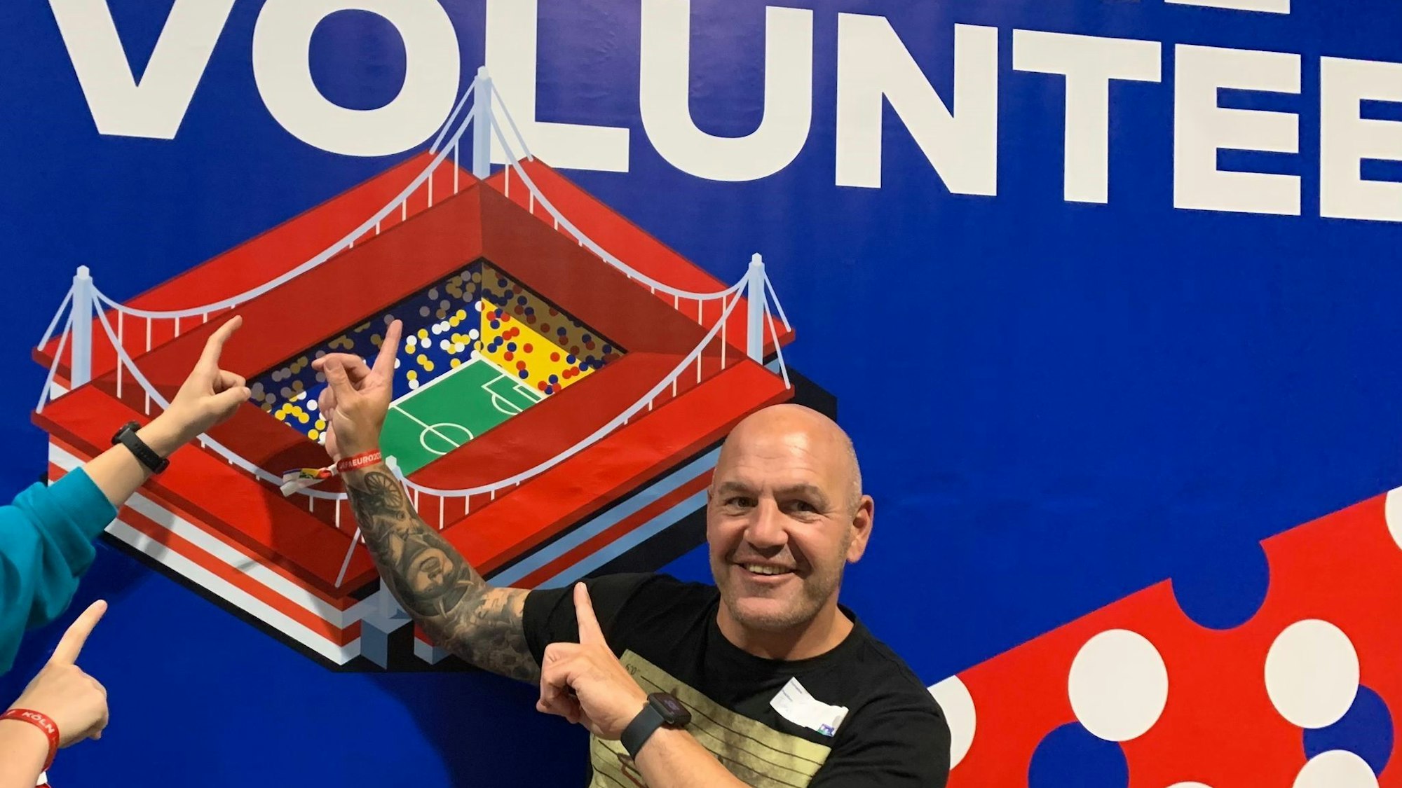 Das Bild zeigt Jörg Piana vor einer Volunteer-Wand in Köln. Er zeigt mit den Fingern auf das Bild des Kölner Stadions.