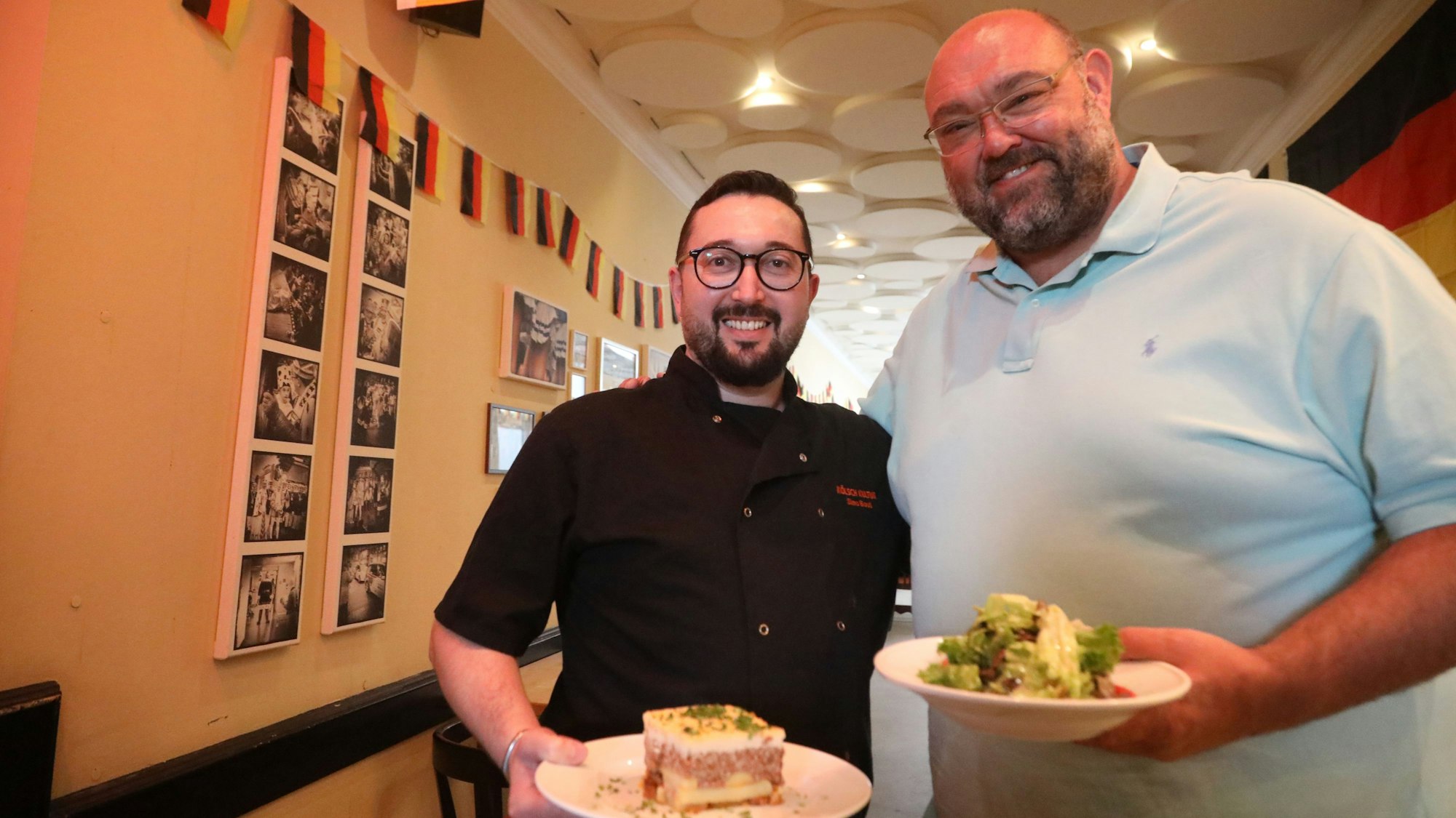 Simo Bouti und Thomas Mick halten einen Teller Moussaka und einen Salat in die Kamera.