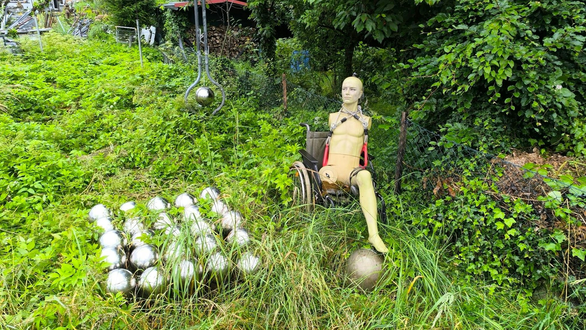 In diesem verwilderten Garten inmitten einer Wohnsiedlung in Leverkusen-Schlebusch sind einige Skulpturen und Figuren zu finden. Dieses Bild zeigt links eine Skulptur, die aus einigen aneinandergefügten Kugeln zusammengesetzt wurde. Rechts sitzt eine Art Schaufensterpuppe in einem Rollstuhl. Die Puppe hat nur ein Bein. Links befindet sich eine Metallkonstruktion aus mehreren Kugeln.
