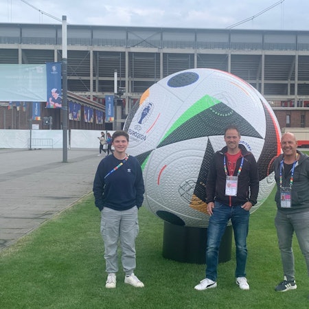 Das Bild zeigt die drei freiwilligen Helfer vor dem Rhein-Energie-Stadion in Köln. Sie stehen vor einem überdimensionalen Spielball.