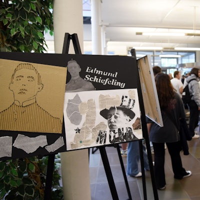 Auf Leinwandständern sind Kunstwerke zu sehen, Menschen stehen zusammen und sprechen.