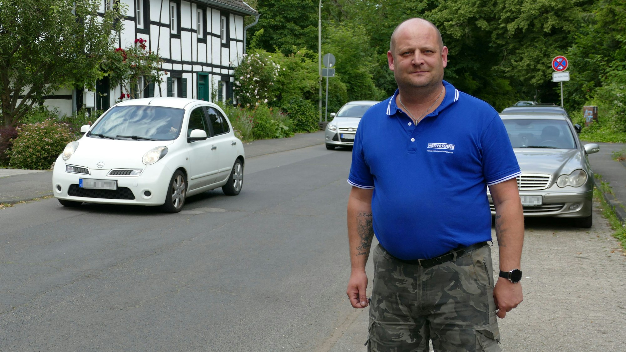 Ein Mann steht auf dem Bürgersteig, hinter ihm parkt ein Wagen, zwei Autos fahren auf der Straße.