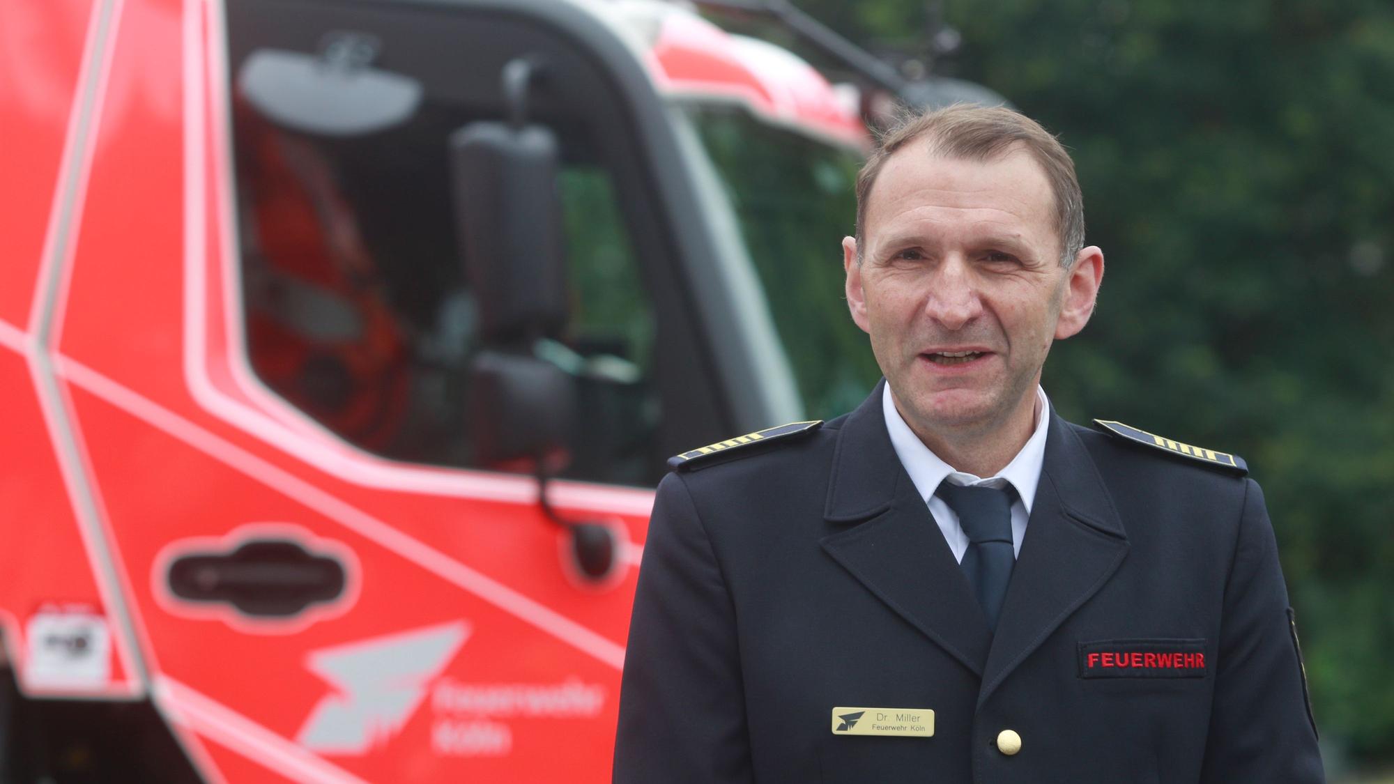 Der Kölner Feuerwehrchef Christian Miller ist im Porträt zu sehen. Er trägt Uniform. Hinter ihm steht ein neues Feuerwehrauto.