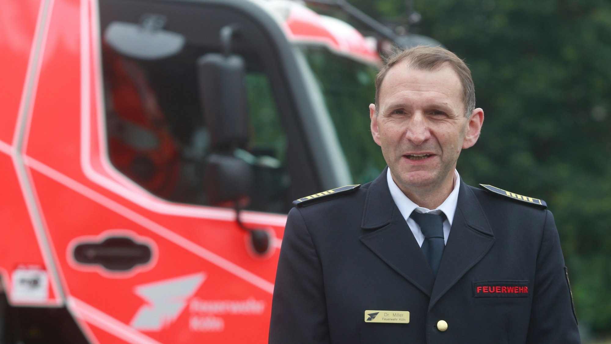 Der Kölner Feuerwehrchef Christian Miller ist im Porträt zu sehen. Er trägt Uniform. Hinter ihm steht ein neues Feuerwehrauto.