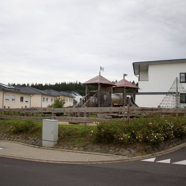 Das Bild zeigt den Spielplatz an der Stotzheimer Straße in Kirspenich von der Straße aus. Im Hintergrund die umliegenden Wohngebäude.