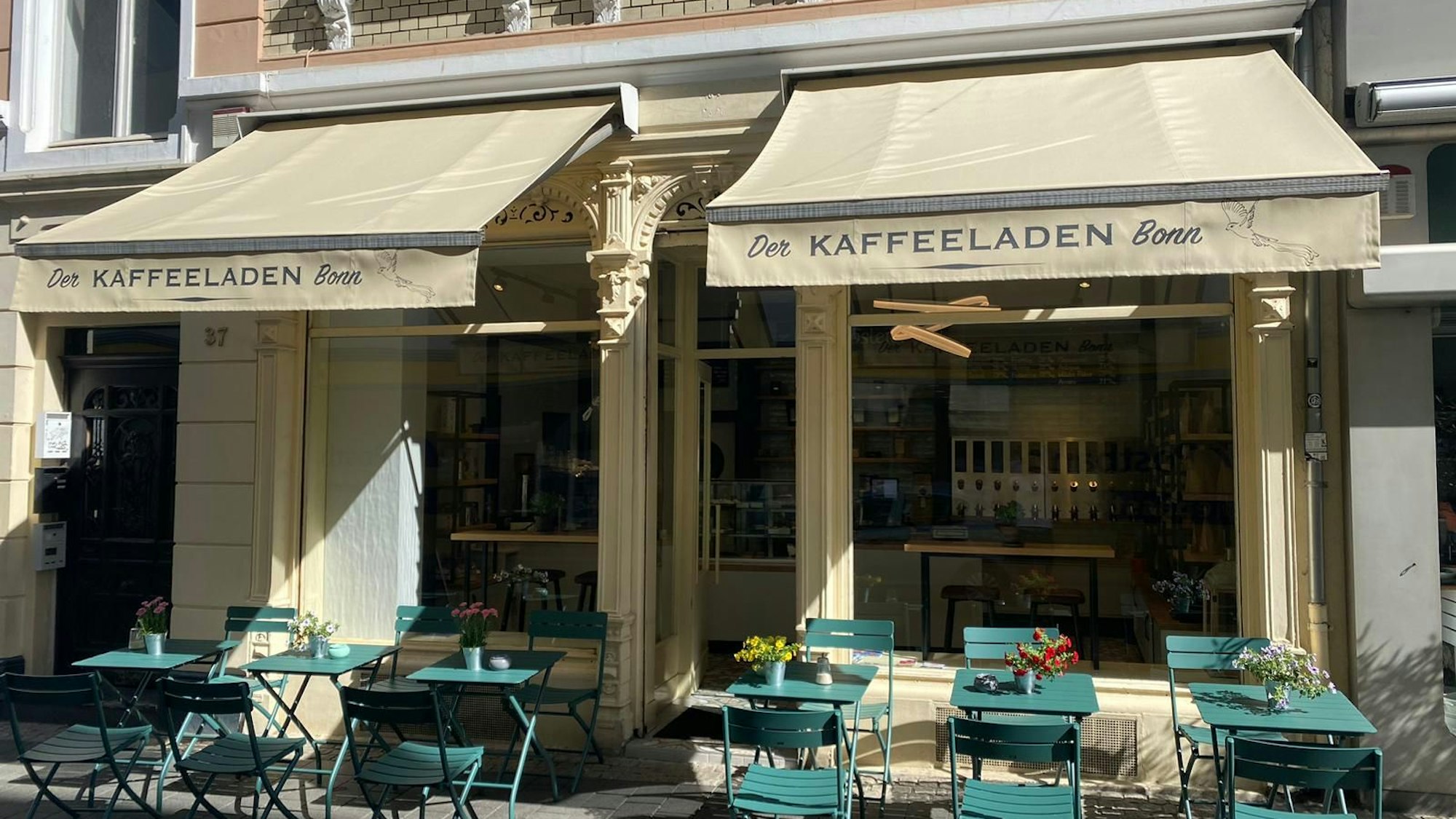 Ein Café mit grünen Stühlen und Tischen im Außenbereich. Zwei Markisen mit der Aufschrift „Der Kaffeeladen Bonn“.