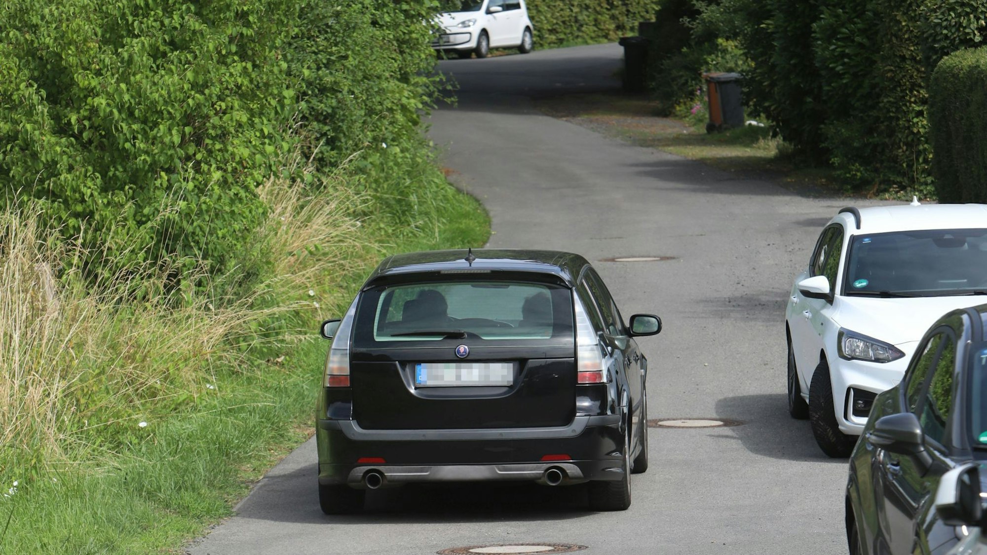 Ein schwarzes Auto fährt eine Straße hinauf, am Fahrbahnrand stehen parkende Fahrzeuge.