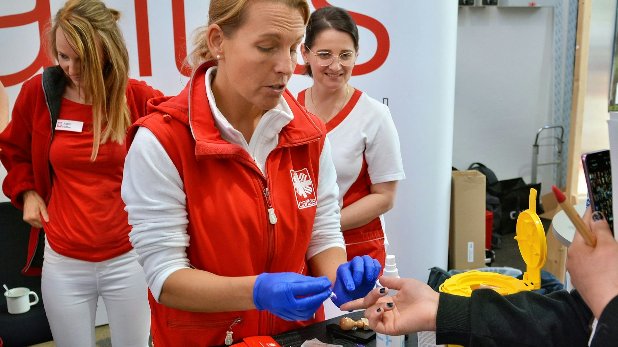 Eine Mitarbeiterin der Caritas, die blaue Einweghandschuhe trägt, nimmt einer Besucherin mit einem Piks in den Finger Blut für eine Untersuchung ab.