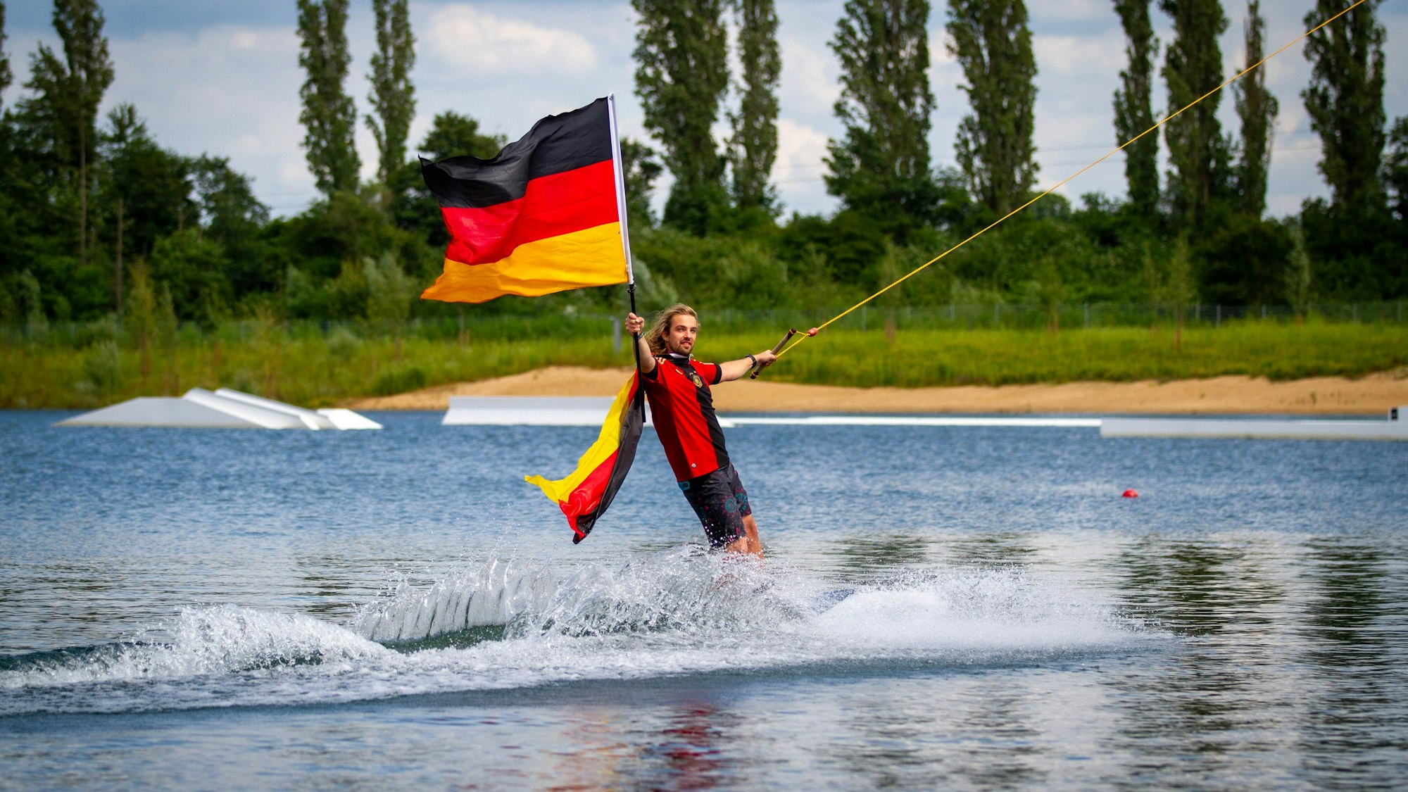 Ein Mann auf einem Wakeboard hält am WakeClubCologne die Deutschlandfahne in die Luft.
