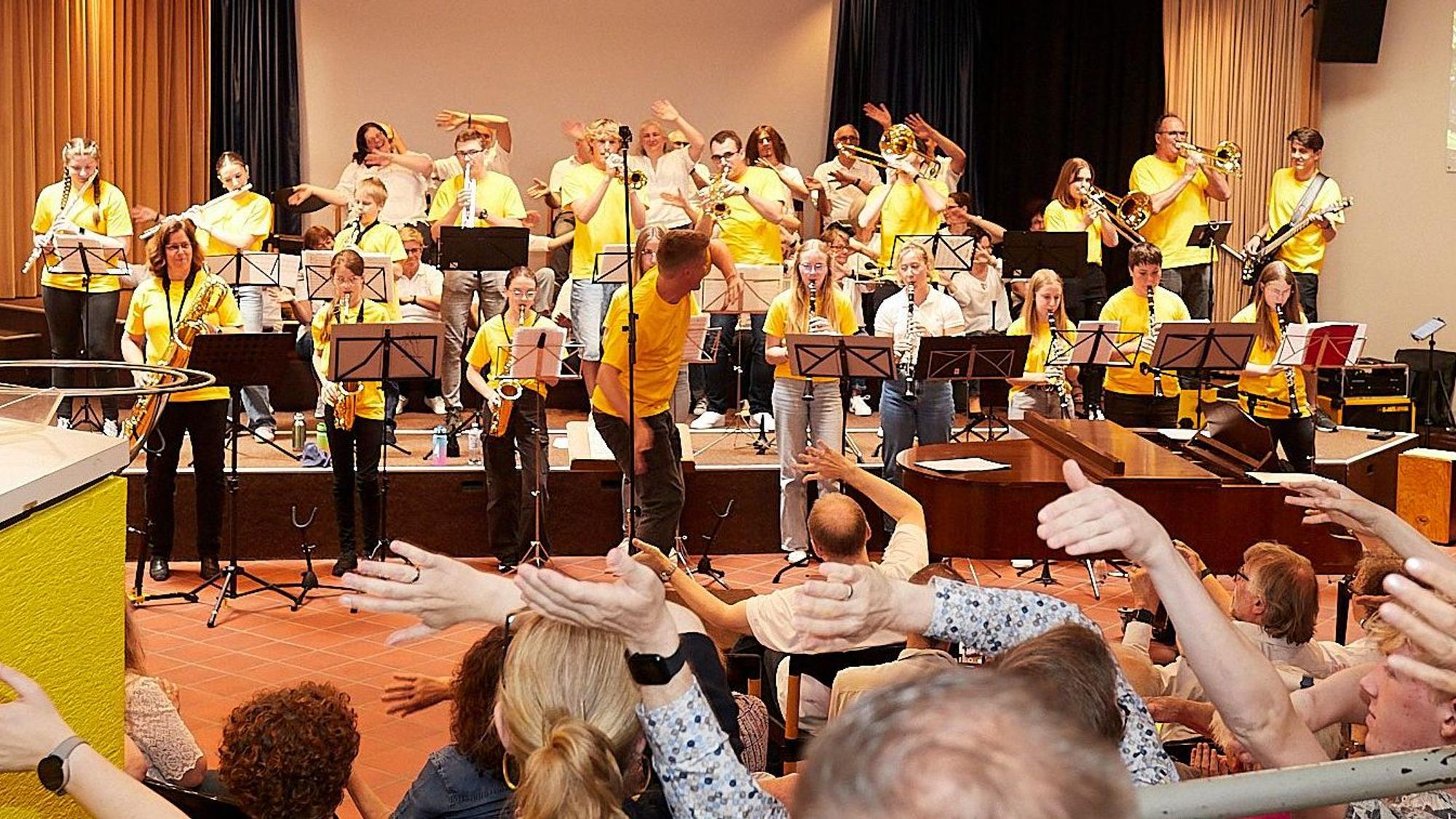 Musiker in gelben Shirts stehen auf der Bühne, das Publikum winkt.