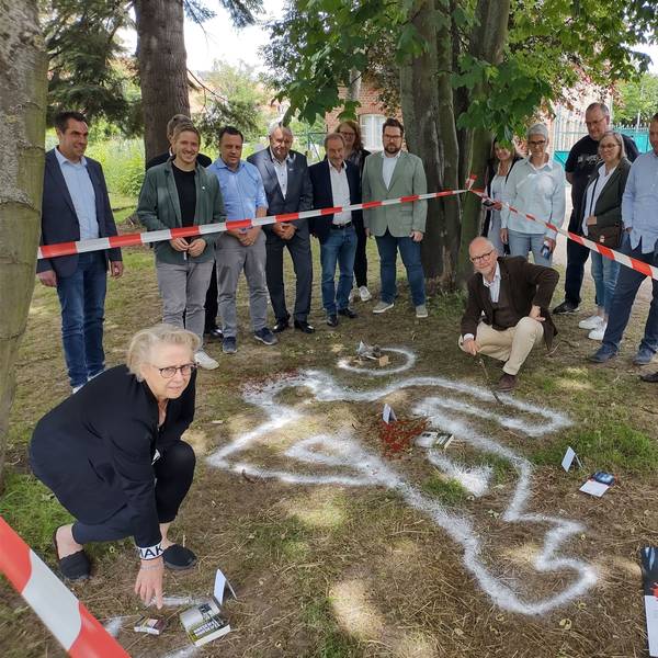 Elke Pistor und Ralf Kramp befinden sich in einem mit Flatterband abgesperrten Bereich, in dem die Umrisse einer imaginären Leiche sowie dargestellte Blutspuren zu sehen sind. Organisatoren und Sponsoren des Krimifestivals schauen ihnen zu.