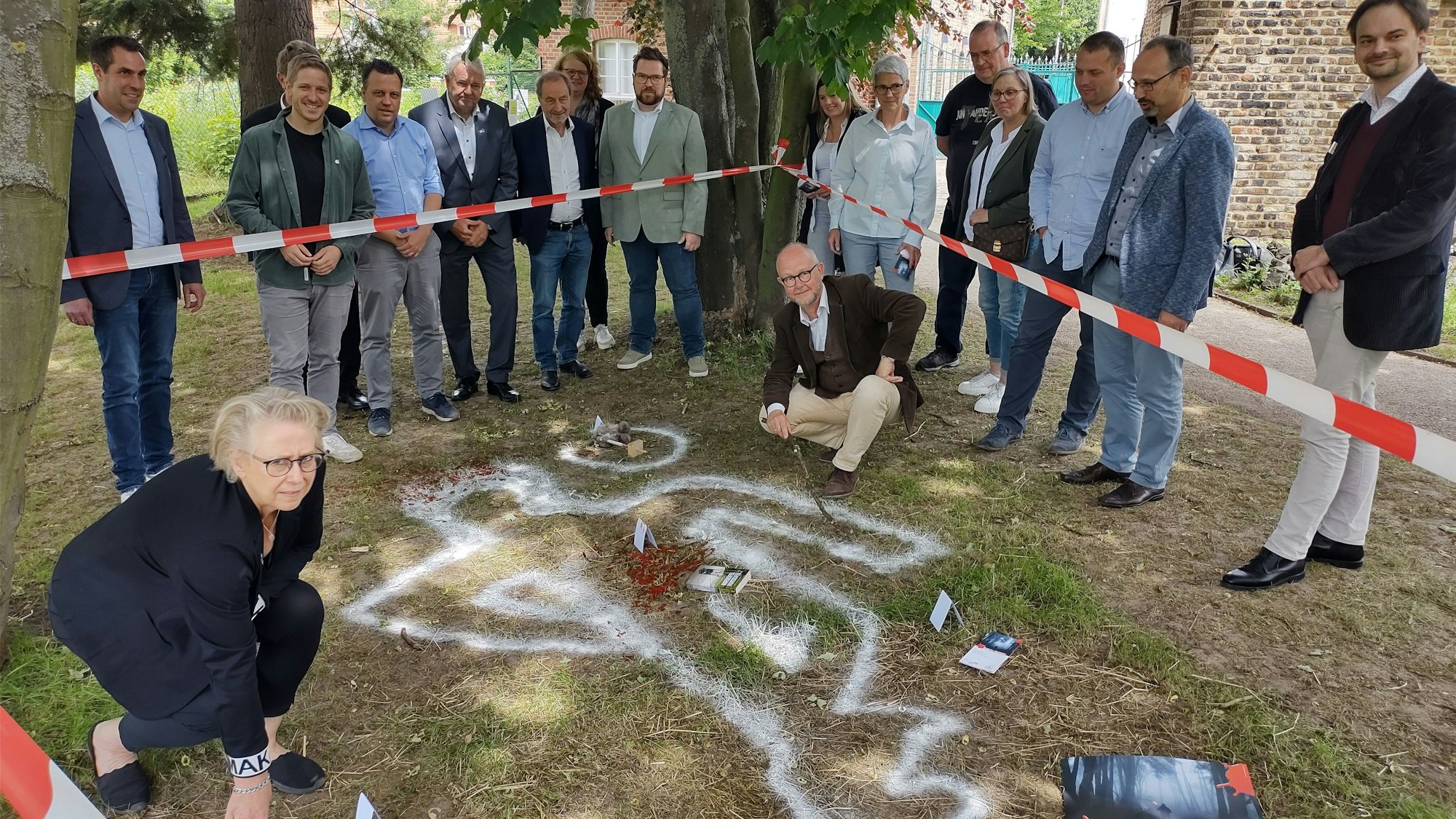 Elke Pistor und Ralf Kramp befinden sich in einem mit Flatterband abgesperrten Bereich, in dem die Umrisse einer imaginären Leiche sowie dargestellte Blutspuren zu sehen sind. Organisatoren und Sponsoren des Krimifestivals schauen ihnen zu.