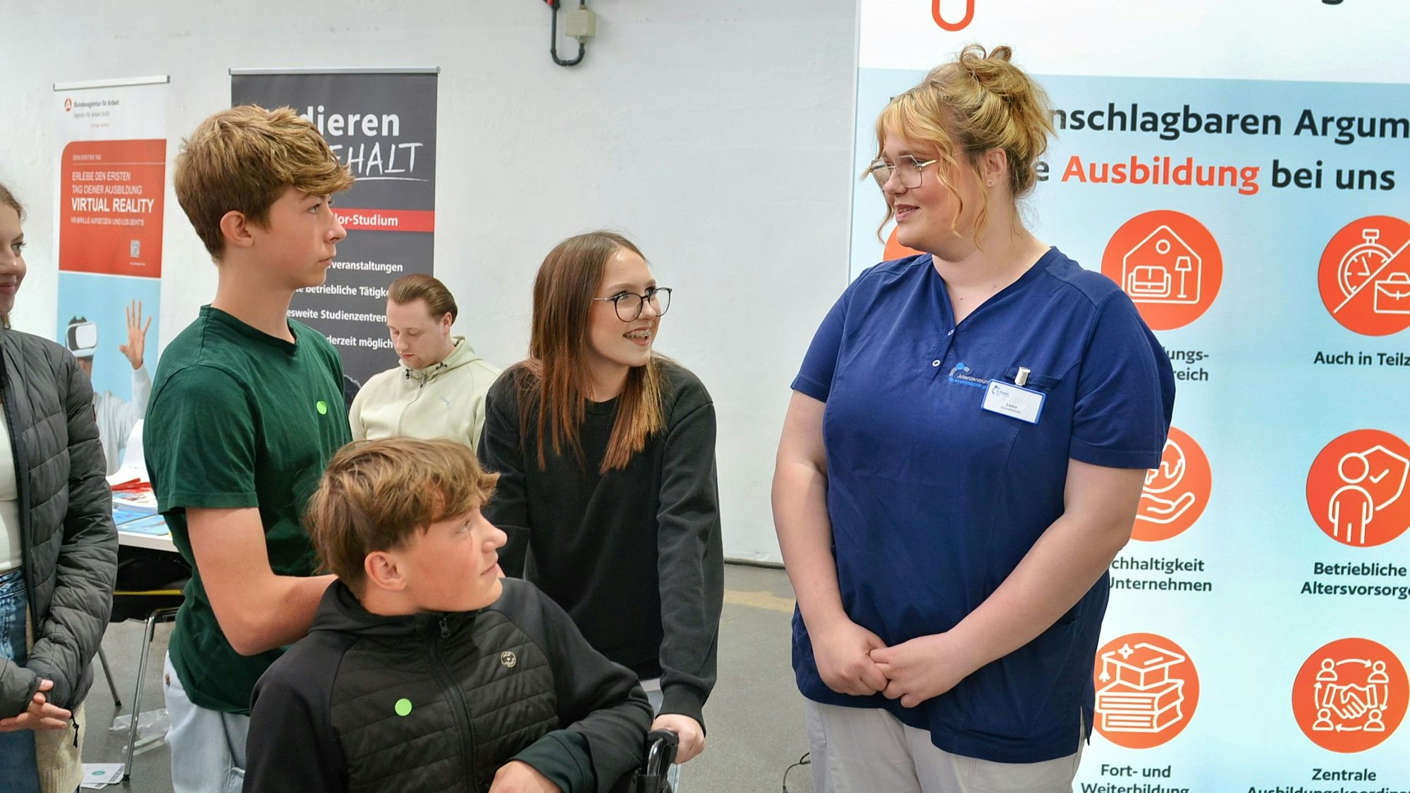 Mehrere Jugendliche sprechen auf einer Berufsmesse mit der Mitarbeiterin eines Pflegedienstes (r.).