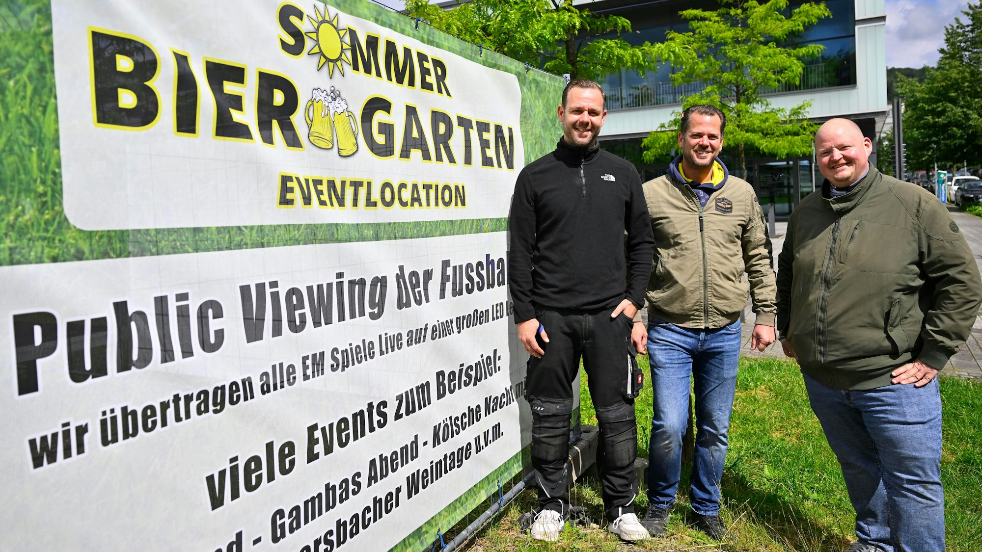 Timo Bay, Frank Hewel und Mike Bierögel stehen vor einem großen Banner, das auch den Sommer Biergarten mit Public-Viewing aller EM-Spiele hinweist.